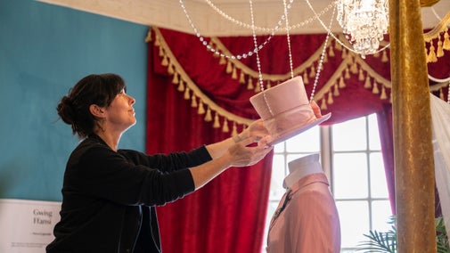Exhibition designer places a pink top hat onto a mannequin, finishing off the costume display.